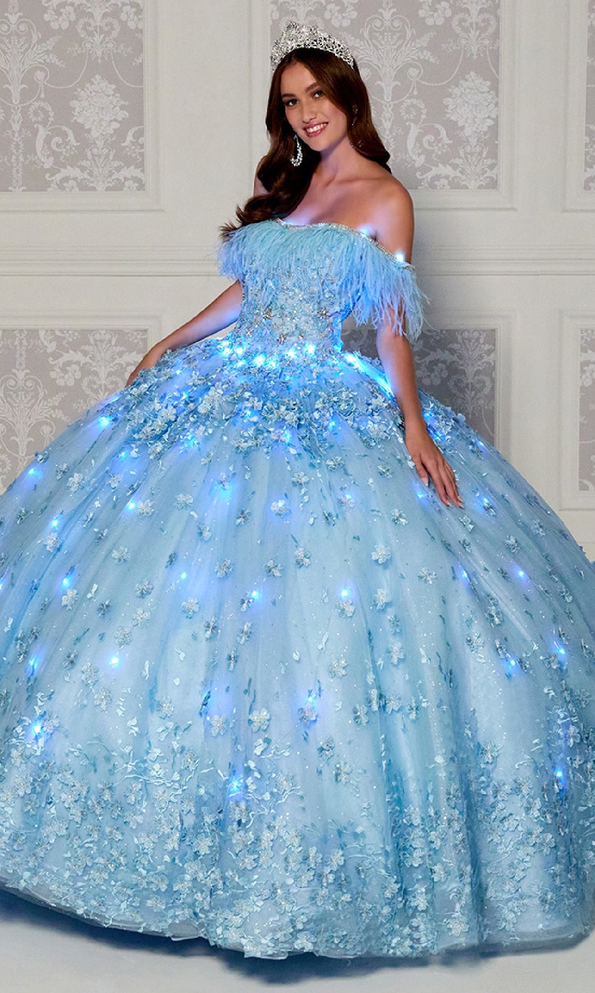 A woman in the Quinceanera Dress PR30115 By Princesa, an off-the-shoulder glowing light blue gown with floral embellishments and a tiara, stands before a patterned wall.